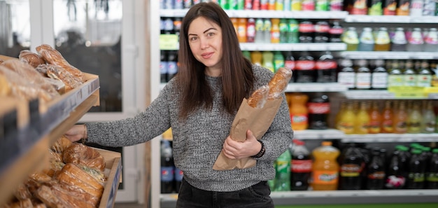 Una bruna sta agli scaffali del negozio sorridente e tenendo il pane di frumento in un sacchetto di carta Look della donna