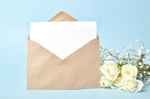 Una busta kraft e un foglio di carta bianco con rose bianche su sfondo blu. Banner per San Valentino. Invito a un matrimonio, anniversario, compleanno