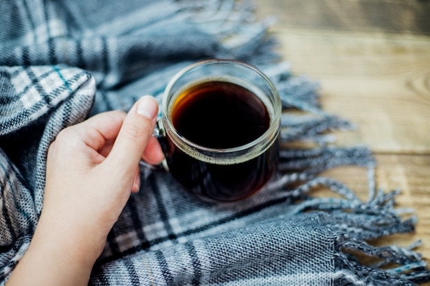 Una calda sciarpa scozzese con frangia giace su uno sfondo di legno chiaro Una tazza di vetro con caffè nero