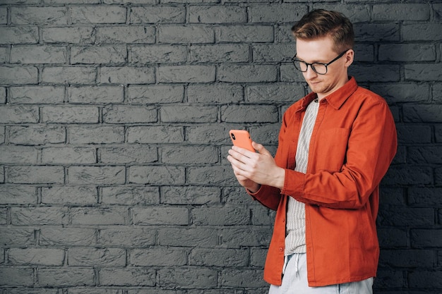 una camicia rossa e occhiali guardando lo smartphone in piedi su uno sfondo di muro di mattoni neri