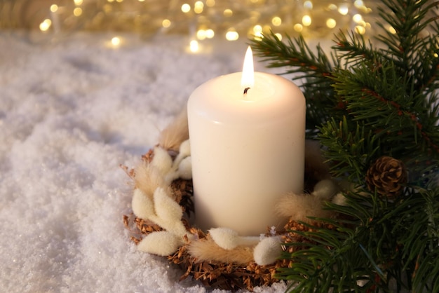 Una candela bianca decorata con decorazioni natalizie sulla neve sullo sfondo delle luci