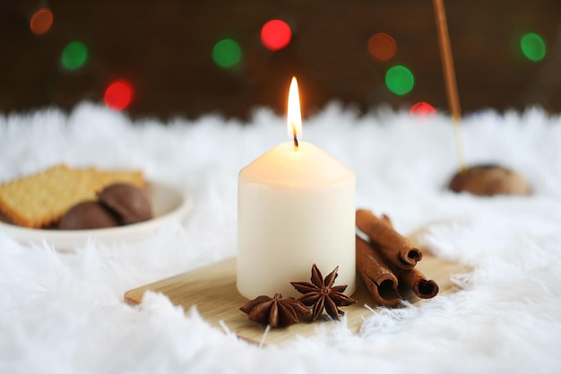 Una candela bianca si trova su una tavola di legno su un plaid bianco Dietro ci sono biscotti e bastoncini di cannella al cioccolato e anice stellato Dietro c'è una ghirlanda di Natale