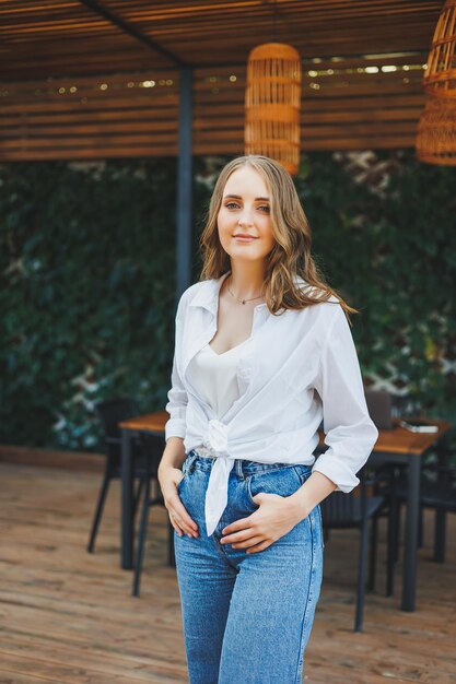 Una carina giovane donna con una camicia bianca e jeans blu è in piedi sulla terrazza di un caffè estivo donna in abiti estivi casuali e eleganti