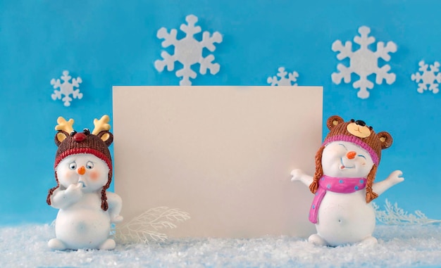 Una cartolina di Natale, un luogo da copiare. Due piccoli giocattoli divertenti del pupazzo di neve in cappelli e sciarpe tricottati nella neve su fondo blu luminoso. Sfondo festivo con pupazzi di neve divertenti. Cartolina d'auguri di felice anno nuovo.