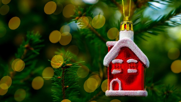 Una casa di Natale rossa è appesa al ramo verde di un albero di Natale con luci bokeh dorate.