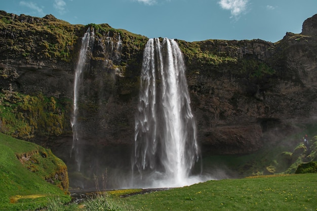 Una cascata in islanda con un campo verde in primo piano e un cielo azzurro con nuvole.