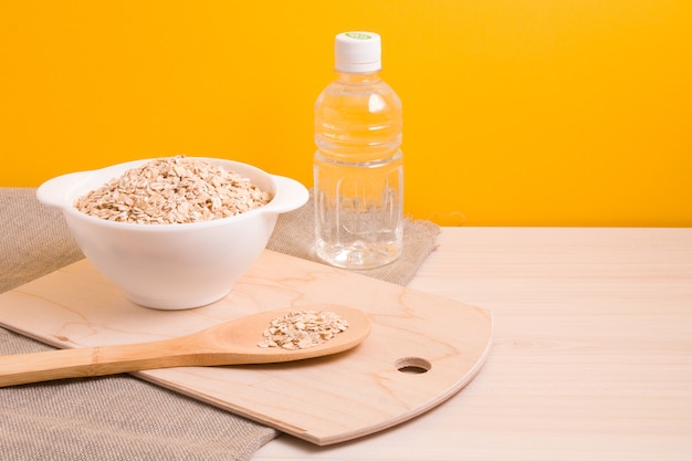 Una ciotola bianca con farina d'avena secca, una bottiglia d'acqua e un grande cucchiaio di legno su un tagliere