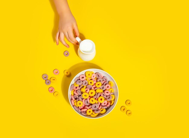 Una ciotola di colorati anelli di cereali su un tavolo giallo Il concetto della colazione Il bambino versa il latte in una ciotola