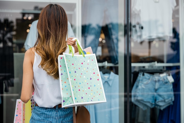 Una cliente felice che porta le borse della spesa sta davanti alla vetrina di un negozio ammirando la mostra di moda. È entusiasta della vendita e pronta a spendere per i suoi stili preferiti.