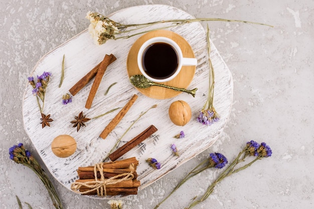 Una composizione di una tazza di caffè nero, bastoncini di cannella, anice e fiori sparsi su un podio di legno bianco e uno sfondo di cemento. vista dall'alto.