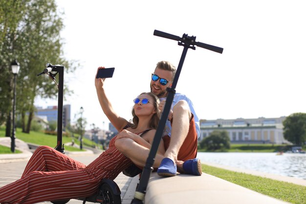 Una coppia adorabile che si diverte a guidare uno scooter elettrico, si prende una pausa dalla guida, si siede sulla sponda del fiume, si fa selfie,