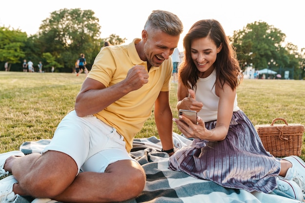 una coppia amorosa adulta emotiva felice all'aperto in un bellissimo parco naturale verde utilizzando il telefono cellulare fa il gesto del vincitore.