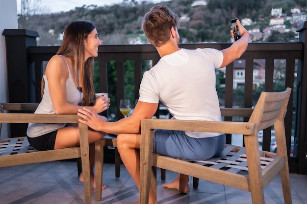 Una coppia caucasica facendo colazione sulla terrazza dell'hotel in pigiama. Fare un selfie con il telefono, con succo d'arancia al mattino, stile di vita di una coppia innamorata