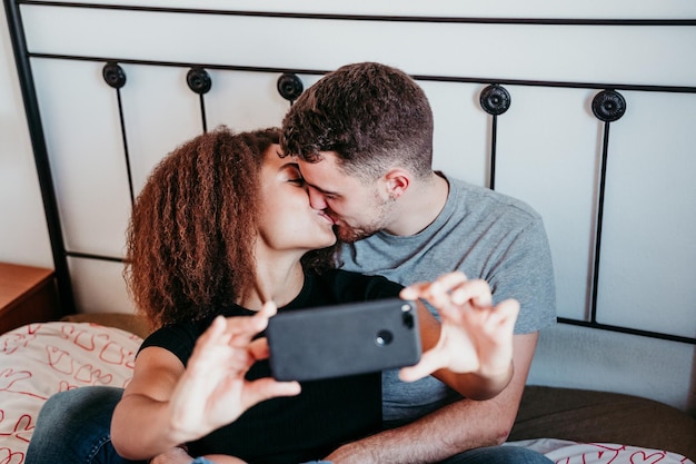 Una coppia che ride seduta sul letto e si bacia mentre si fa un selfie