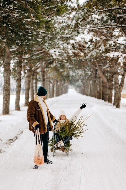 Una coppia che si diverte con la slitta in una foresta di alberi di Natale, mangiando mandarini e godendosi una neve