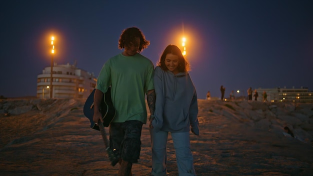 Una coppia di adolescenti rilassati che camminano la sera sulla spiaggia sabbiosa un musicista con la chitarra in mano