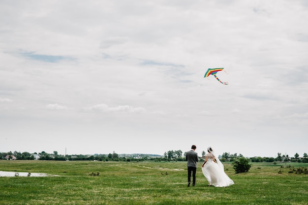 Una coppia di sposi correre indietro intorno al campo lasciando volare l'aquilone e tenere l'aquilone Sposa e sposo a piedi.