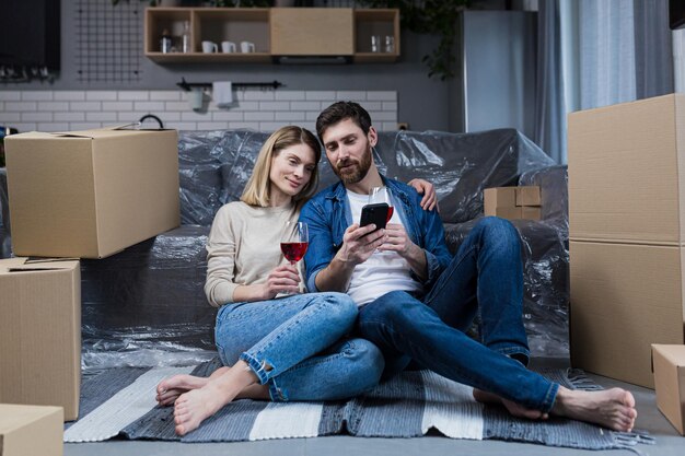 Una coppia di sposi innamorati, un uomo e una donna celebrano una festa di inaugurazione della casa, bevono vino dalle scatole di cartone e scelgono nuovi mobili ed elettrodomestici nel negozio online usa il telefono