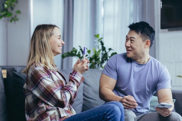 Una coppia felice di uomini e donne di razze diverse si rallegra del risultato del test di gravidanza giovane famiglia a casa seduta sul divano e abbracciata