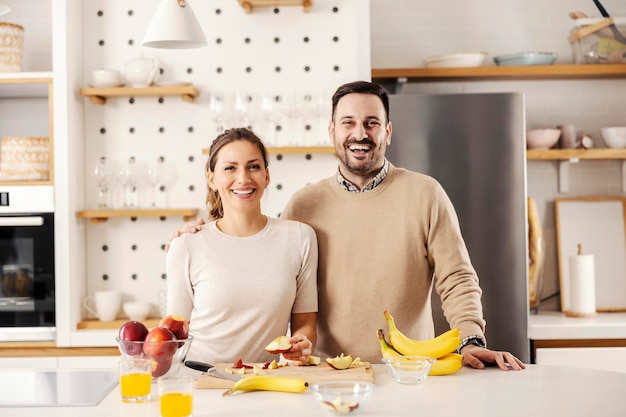 Una coppia felice in cucina prepara frutta e sorride alla telecamera a casa loro