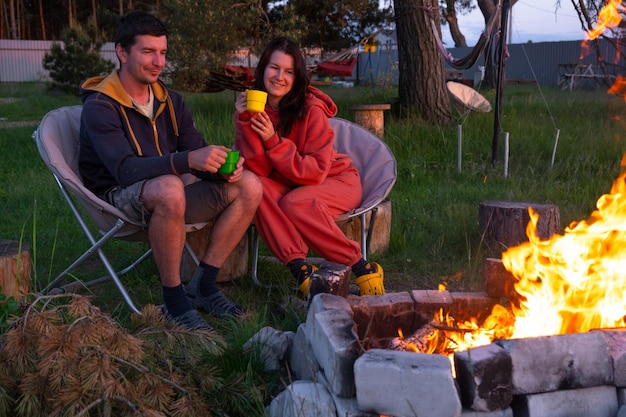 Una coppia innamorata è seduta vicino al fuoco all'aperto nel cortile della casa su sedie da campeggio una sera romantica un uomo e una donna si scaldano le mani vicino al fuoco bevono tè