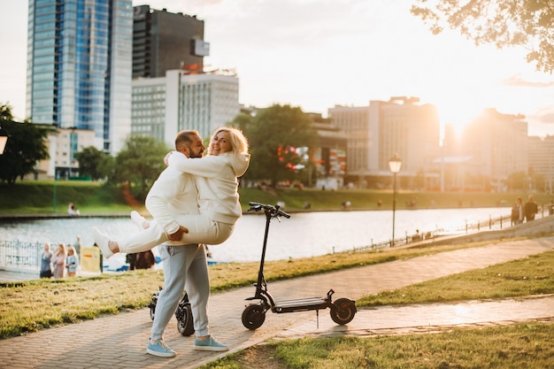 Una coppia innamorata in abiti bianchi si trova in città al tramonto vicino a uno scooter elettrico.