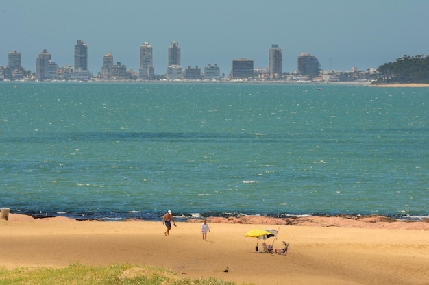 Una coppia passeggia sulla spiaggia mentre sullo sfondo si intravede lo skyline di Punta del Este