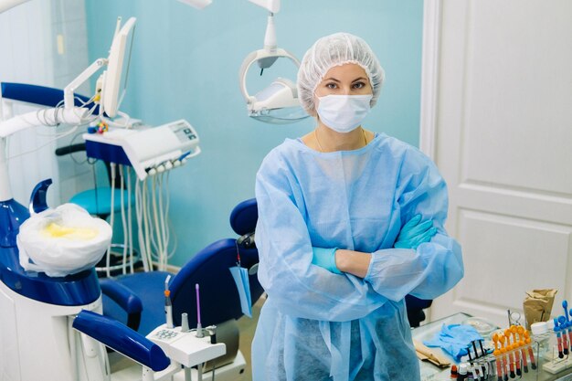 Una dentista donna che indossa una maschera medica e guanti di gomma posa per la fotocamera e piega le braccia nel suo ufficio