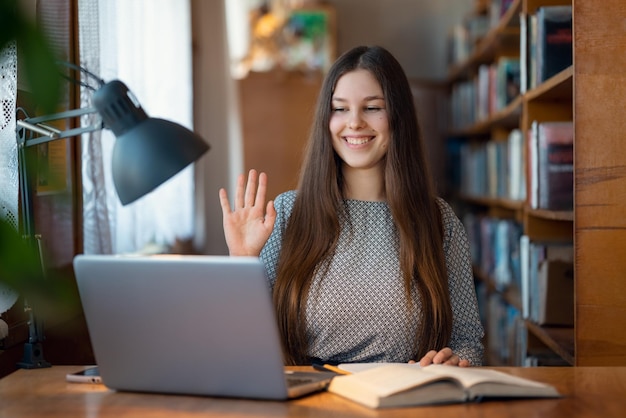 Una diligente studentessa universitaria che tiene lezioni online, dai saluti della biblioteca con i compagni di classe alla webcam del suo laptop.