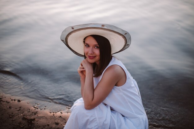 Una dolce e sofisticata giovane donna in abito bianco e cappello di paglia è accovacciata in riva al fiume o al mare...