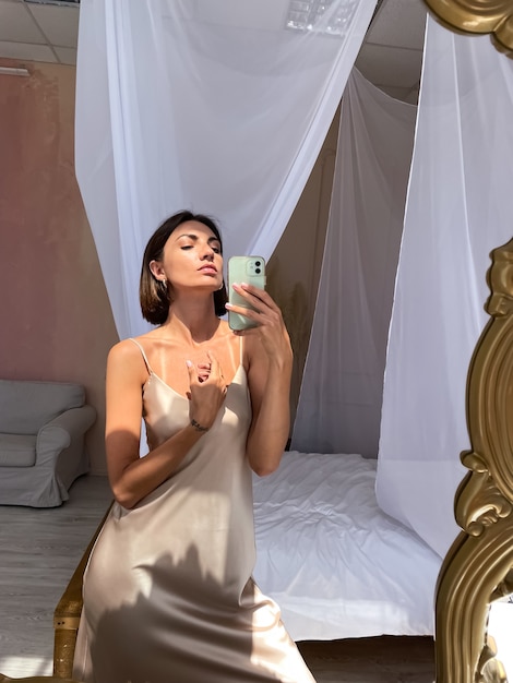 Una donna abbronzata in forma in un romantico abito di seta beige a casa scatta una foto selfie al telefono allo specchio