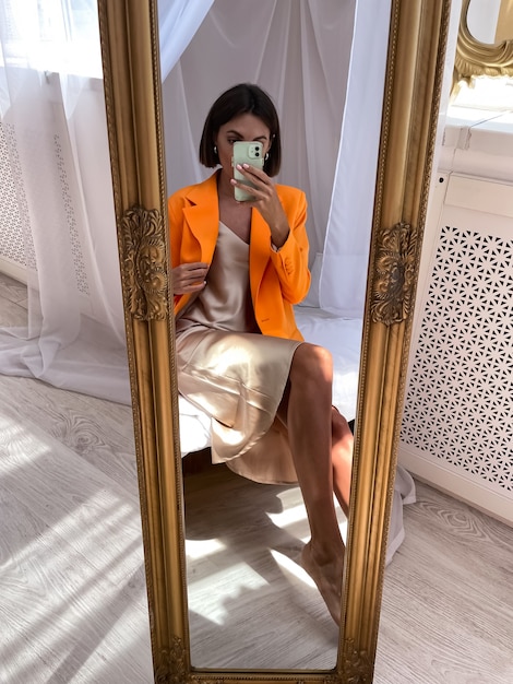 Una donna abbronzata in forma in un romantico abito di seta beige e un blazer arancione a casa scatta una foto selfie al telefono allo specchio