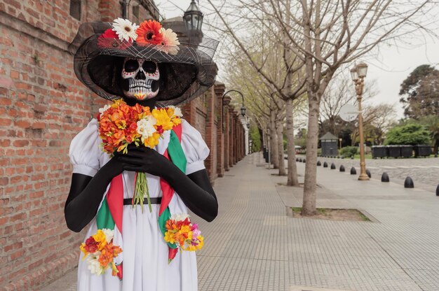 Una donna adulta con il costume di Catrina e il trucco in piedi con gli occhi chiusi e con in mano dei fiori