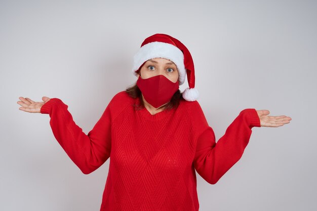 Una donna adulta con una maschera protettiva rossa, un cappello da Babbo Natale su uno sfondo grigio, allargò le braccia per la sorpresa. Natale, Capodanno in quarantena.