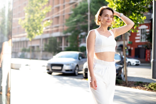 Una donna adulta di successo in abito bianco cammina lungo una strada illuminata e si raddrizza i capelli sullo sfondo di una casa in costruzione e di automobili. giornata mondiale del turismo