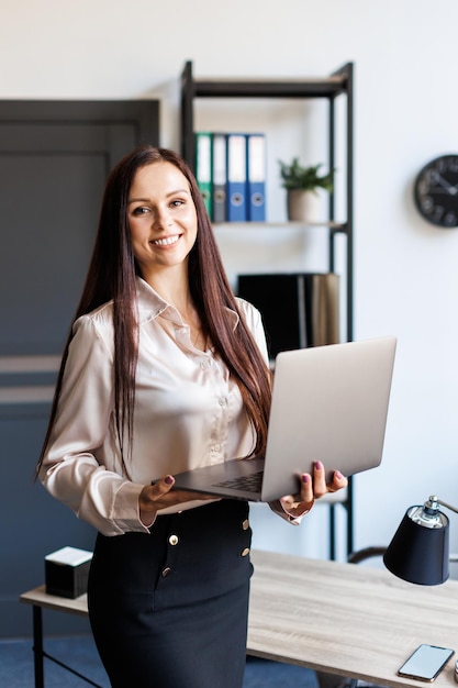 Una donna adulta di successo in piedi in un ufficio moderno tiene in braccio un computer portatile Felice donna d'affari brunettehair in abito elegante ed elegante guarda la fotocamera e sorride