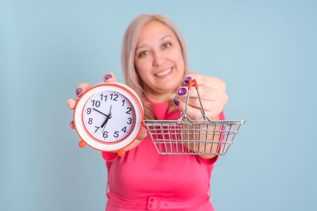 Una donna adulta sorridente tiene un orologio e un piccolo carrello della spesa a distanza di un braccio, messa a fuoco selettiva