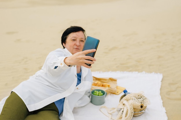 Una donna adulta sulla spiaggia vicino all'oceano in primavera durante un picnic fa un selfie parla di un video