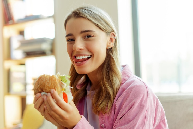 Foto una donna affamata e sorridente lecca le labbra mangiando un hamburger guardando la telecamera seduta in un caffè moderno