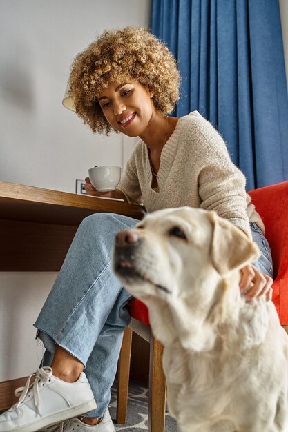 Una donna afroamericana felice e ricca si diverte a bere caffè vicino al Labrador in un hotel per animali domestici.