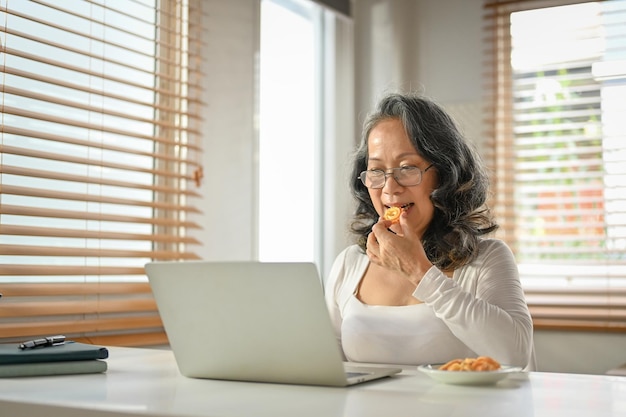 Una donna agghiacciante degli anni '60 che mangia biscotti mentre usa il suo laptop nel suo spazio di lavoro domestico