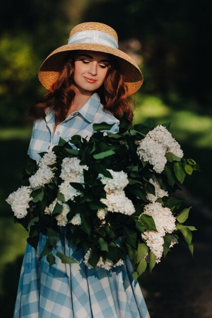 Una donna alla moda in un cappello di paglia posa sui fiori lilla in un soleggiato parco primaverile