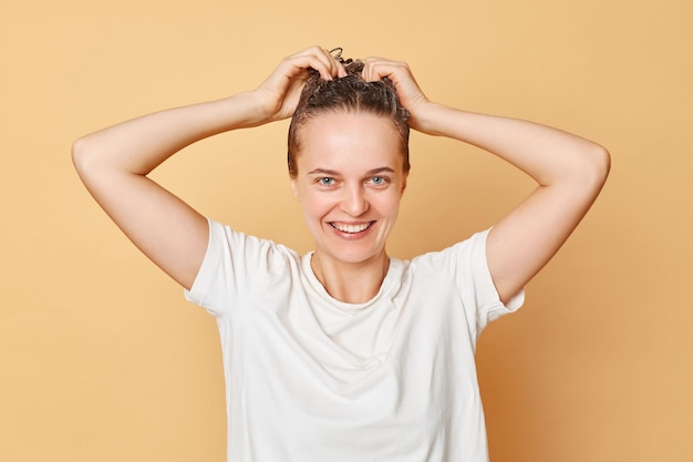 Una donna allegra che indossa una maglietta bianca lava i capelli in piedi con schiuma di shampoo sulla testa isolata su sfondo beige facendo un bagno rilassante per coccolarsi