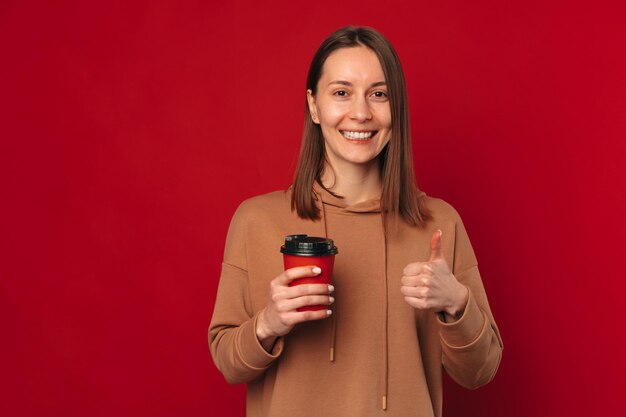 Una donna allegra consiglia questo caffè bianco con in mano una tazza da asporto e il pollice alzato