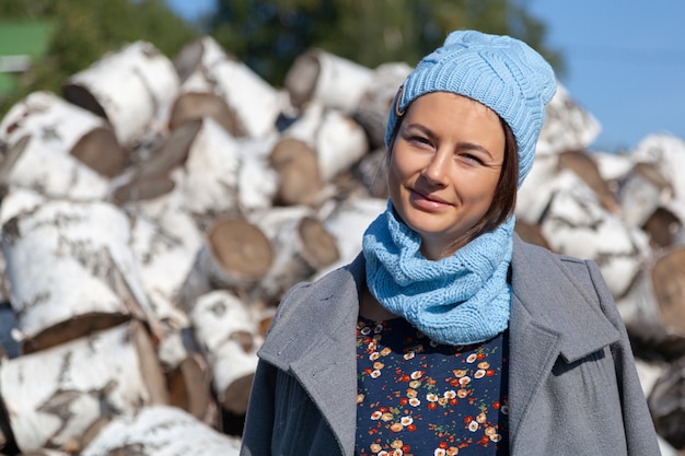 Una donna allegra e attraente in abiti eleganti (un abito con motivi floreali, un cappello lavorato a maglia e un cappotto) sorride, gode della natura e si siede su una montagna di tronchi di betulla. Concetto di vita del villaggio