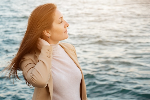 Una donna allegra tocca i capelli lunghi in piedi sul lungomare godendosi la bellissima natura contro la vista ravvicinata estrema del mare increspato