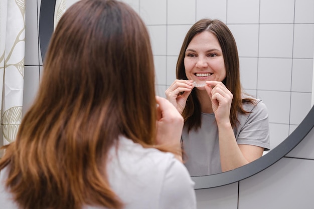 Una donna ampiamente sorridente davanti a uno specchio in bagno con in mano allineatori di denti di plastica invisibili Indossare parentesi graffe Sorriso bello e sano