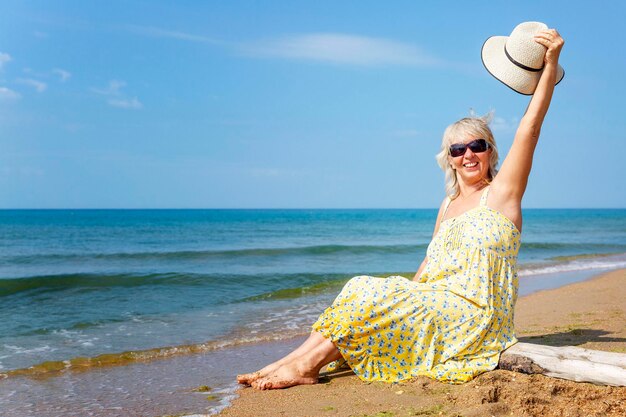 Una donna anziana che ride con un vestito giallo e un cappello si sta riposando sulla riva in una giornata di sole Vacanze al resort e viaggi