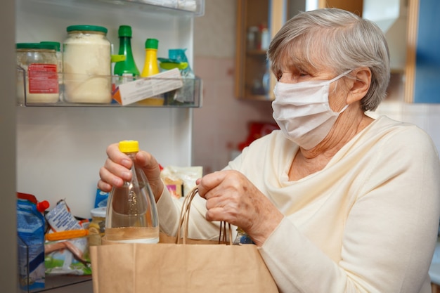 Una donna anziana con una mascherina medica prende il cibo dai pacchetti e lo mette in frigorifero