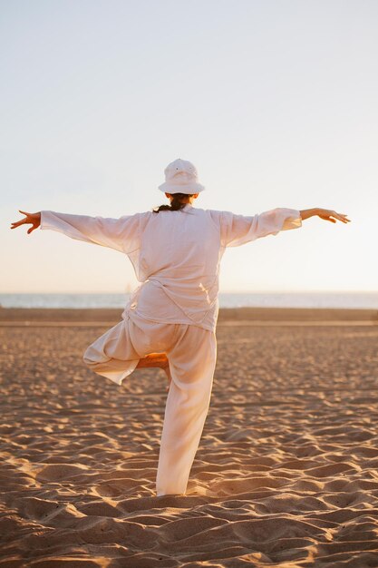 Una donna anziana di oltre 50 anni vestita di bianco sta facendo yoga su una spiaggia